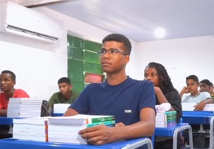 Estudante de óculos sentado em sala de aula, com pilhas de livros sobre a carteira e colegas ao fundo.