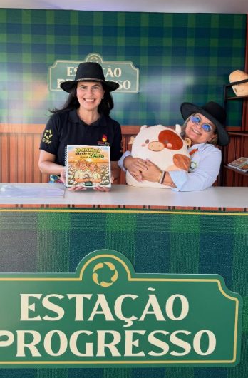 Duas mulheres de chapéu no balcão da loja itinerante Estação Progresso, uma segurando um livro infantil e a outra abraçando uma pelúcia de vaquinha.