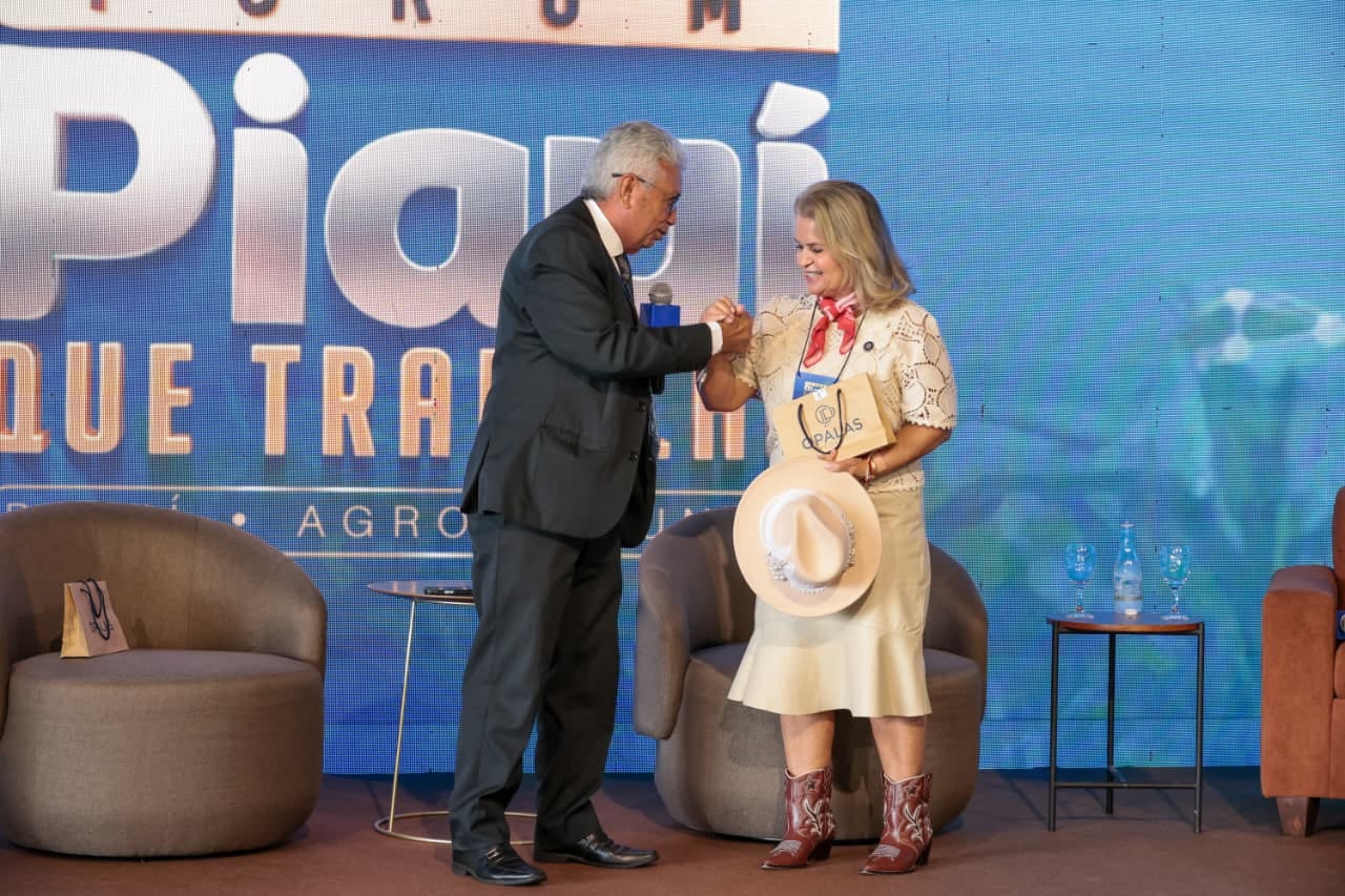 Ani Sanders é cumprimentada no palco, segurando um chapéu e uma sacola de presente.