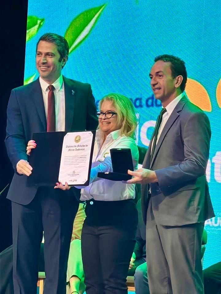 Ani Sanders recebe a medalha e o diploma no palco, entre duas autoridades.