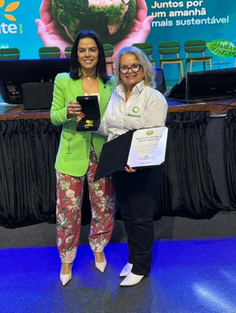 Ani Sanders ao lado de uma convidada, as duas segurando a medalha e o diploma.